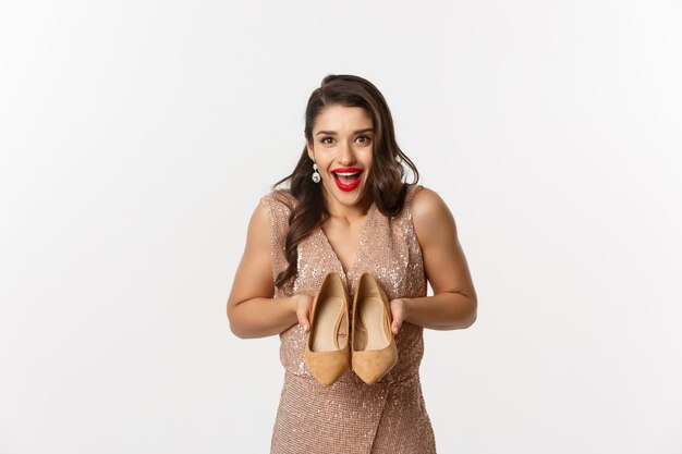 Concepto de fiesta y celebración. Mujer emocionada mostrando nuevo par de tacones y sonriendo, con un vestido elegante, de pie sobre fondo blanco.