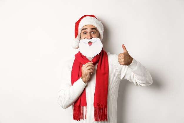 Concepto de fiesta y celebración de Navidad. Feliz modelo masculino con sombrero de Santa Claus y máscara de barba blanca, mostrando el pulgar hacia arriba gesto, de pie sobre fondo blanco.