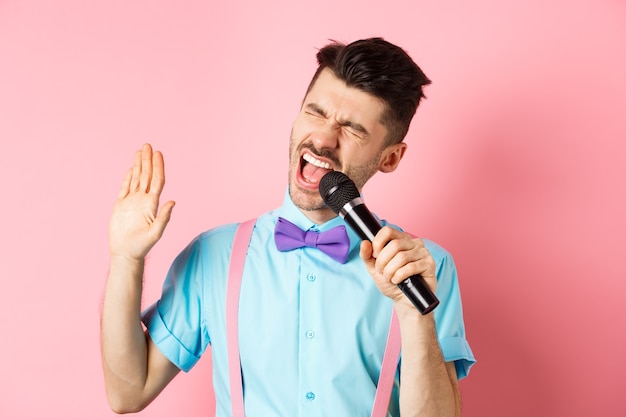 Concepto de fiesta y eventos festivos. Chico divertido realiza una canción, canta karaoke en micrófono con cara despreocupada, de pie con pajarita y tirantes sobre fondo rosa.