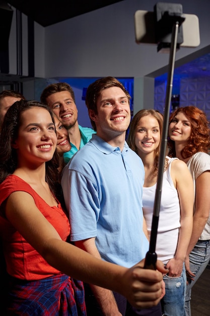 concepto de fiesta, tecnología, vida nocturna y personas - amigos sonrientes con smartphone y monopod tomando selfie en el club