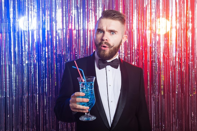 Concepto de fiesta y vacaciones de club - retrato de hombre guapo elegante con un traje caro tiene cóctel azul sobre fondo brillante.