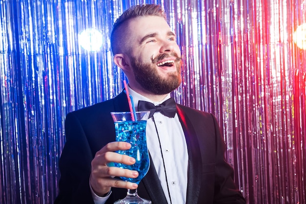 Concepto de fiesta y vacaciones de club - retrato de hombre guapo feliz elegante en un traje caro tiene cóctel azul sobre fondo brillante.