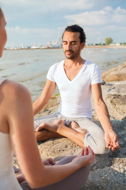 concepto de fitness, deporte, amistad y estilo de vida - pareja sonriente haciendo ejercicios de yoga sentados al aire libre