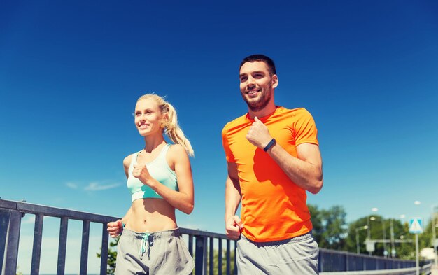 Foto concepto de fitness, deporte, amistad y estilo de vida saludable - pareja sonriente con reloj de frecuencia cardíaca corriendo en la playa de verano