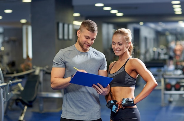 concepto de fitness, deporte, ejercicio y dieta - mujer joven sonriente y entrenadora personal con plan de ejercicios de escritura de portapapeles en el gimnasio