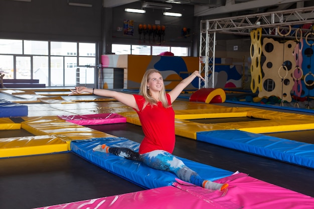 Concepto de fitness, deporte, ejercicio, estiramiento y personas - mujer sonriente haciendo divisiones en la estera sobre fondo de gimnasio.