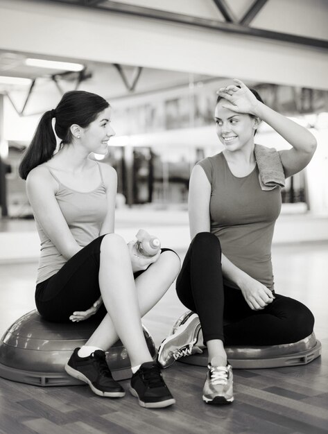 concepto de fitness, deporte, entrenamiento, gimnasio y estilo de vida: dos mujeres sonrientes sentadas en la media pelota y relajándose después de clase en el gimnasio