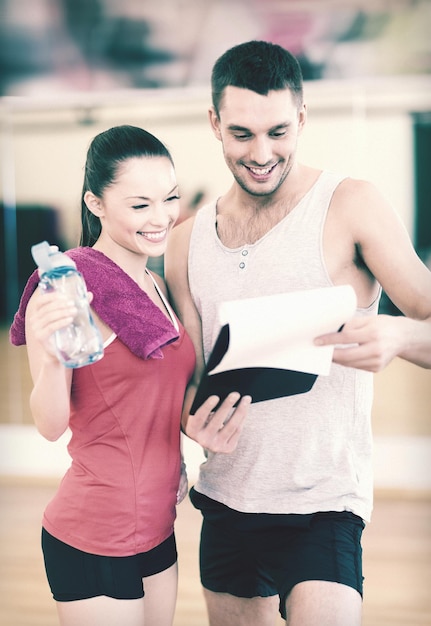 concepto de fitness, deporte, entrenamiento, gimnasio y estilo de vida: entrenador masculino sonriente con portapapeles y mujer con botella de agua en el gimnasio