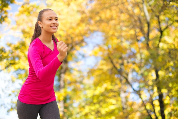 concepto de fitness, deporte, entrenamiento y personas - mujer afroamericana sonriente corriendo al aire libre sobre el fondo de los árboles de otoño