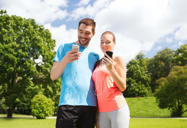 concepto de fitness, deporte, entrenamiento, tecnología y estilo de vida - dos personas sonrientes con smartphones y auriculares al aire libre