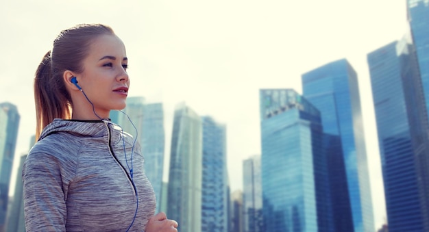 concepto de fitness, deporte, gente, tecnología y estilo de vida - mujer feliz corriendo y escuchando música en auriculares sobre el fondo de los rascacielos de la ciudad de Singapur