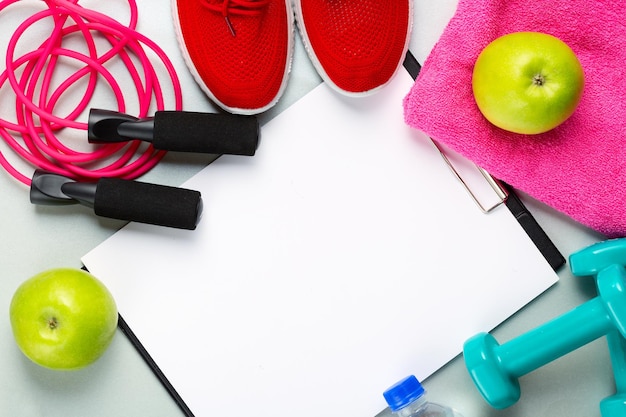 Concepto de fitness y deporte. Zapatillas rojas, manzanas, saltar la cuerda, pesas, botella de agua, toalla rosa y hoja de papel en blanco sobre fondo claro.