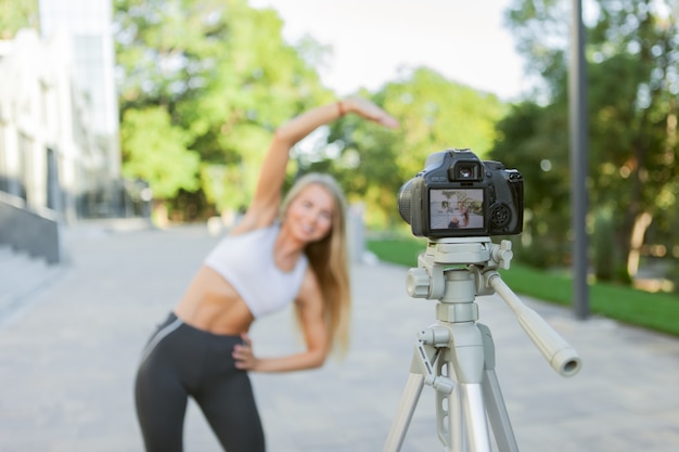 Concepto de fitness, deportes y blogs de vídeo. Blogger de deporte femenino con cámara en trípode grabando entrenamiento físico al aire libre