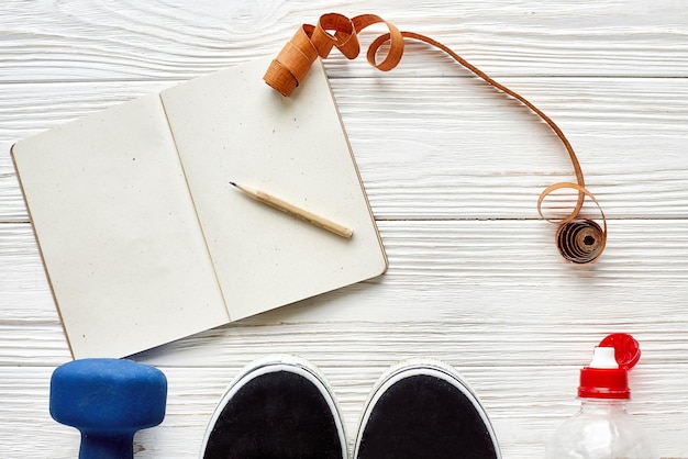 Foto concepto de fitness con lápiz de cuaderno para objetivos deportivos y resoluciones pesas y zapatillas de deporte y cinta de centímetro de botella de agua en el espacio de fondo de madera blanca para texto plano estilo de vida saludable