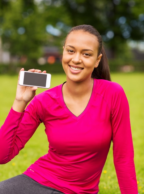 concepto de fitness, parque, tecnología y deporte - mujer afroamericana sonriente mostrando smartphone al aire libre