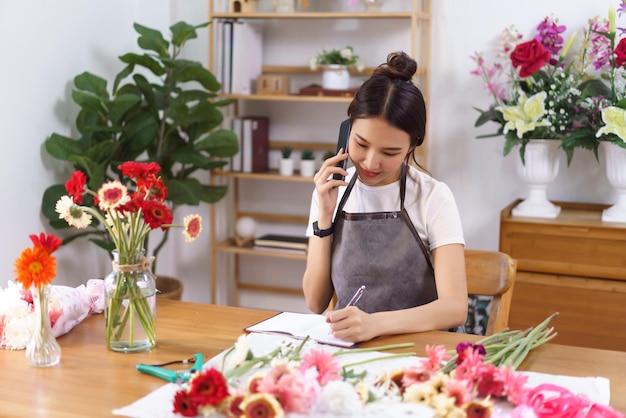 Concepto de floristería Floristería femenina hablando con el cliente por teléfono y tomando nota en el cuaderno en la tienda
