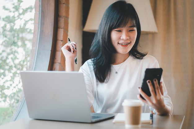 Un concepto de freelance de negocios Una joven asiática sonriente con aparatos ortopédicos trabajando en una computadora portátil y revisando su teléfono inteligente en un espacio de trabajo acogedor