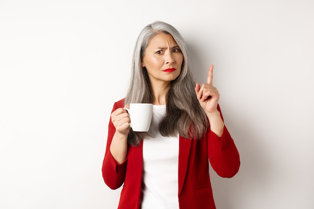 Concepto de gente de negocios. La empresaria asiática disgustada bebiendo café y agitando el dedo con desaprobación, prohíbe algo, de pie sobre fondo blanco.