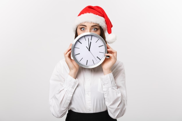 Concepto de gestión del tiempo - mujer de negocios joven con sombrero de santa escondido detrás de un reloj aislado sobre una pared blanca.