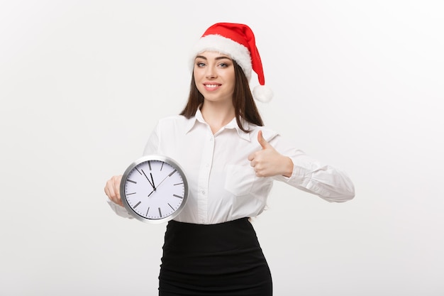 Concepto de gestión del tiempo - mujer de negocios joven con sombrero de santa sosteniendo un reloj y mostrando un golpe aislado sobre una pared blanca.