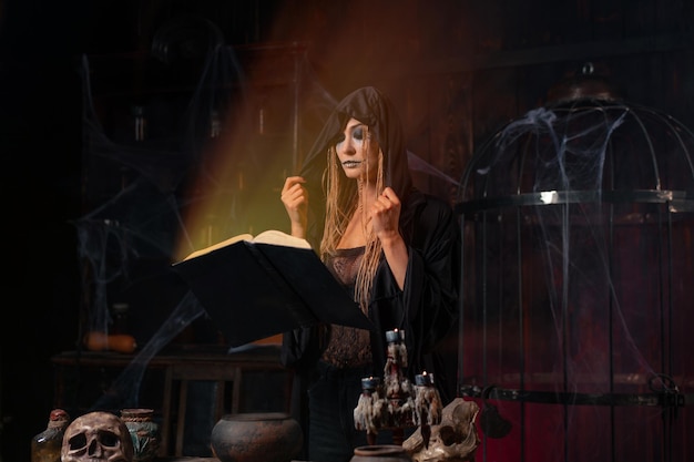 concepto de Halloween. Bruja vestida con capucha negra de pie en la habitación de la mazmorra oscura usa el libro mágico ve la luz del libro conjurando el hechizo mágico. Habitación gótica del mago nigromante femenino con calavera, jaula, telaraña