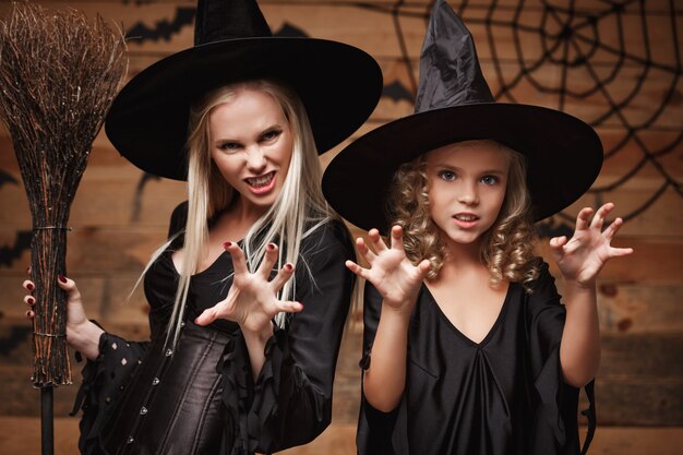 Concepto de Halloween - Closeup madre caucásica y su hija en disfraces de brujas celebrando Halloween posando con calabazas curvas sobre murciélagos y telaraña en la pared de madera.