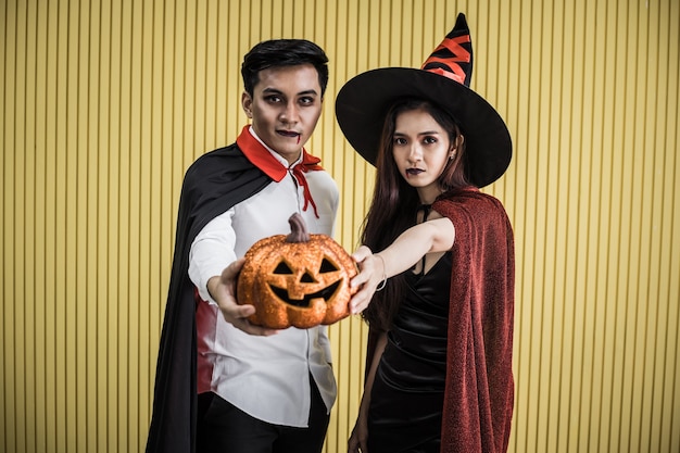 Concepto de Halloween de joven asiática en traje de bruja y hombre asiático en traje de Drácula sobre fondo amarillo. Retrato de pareja de adolescentes disfrazados de brujas y Drácula para celebrar el festival de Halloween.