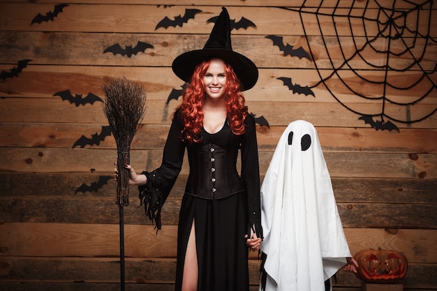 Concepto de Halloween - madre de bruja y pequeño fantasma blanco haciendo truco o trato celebrando Halloween posando con calabazas curvas sobre murciélagos y tela de araña en el fondo de madera del estudio.