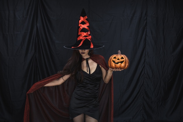 Concepto de Halloween de mujer asiática joven en traje de bruja y mantenga calabaza de Halloween naranja sobre fondo de tela negra. Retrato de mujer adolescente vestida como bruja para celebrar el festival de Halloween.