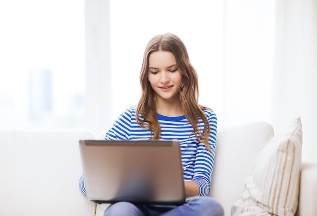 concepto de hogar, tecnología e Internet - adolescente sonriente tendida en el sofá con computadora portátil en casa