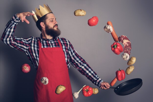 Foto concepto de hombre de cocina, rey de la cocina, hombre barbudo en camisa a cuadros, soltar carne y verduras de una sartén, foto de estudio sobre fondo gris