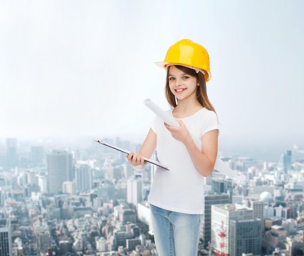 concepto de infancia, construcción, arquitectura, construcción y personas - niña sonriente con casco protector con portapapeles y plano