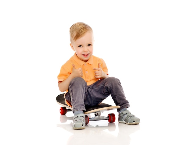 concepto de infancia, deporte, ocio, gestos y personas - niño pequeño feliz sentado en patineta y mostrando los pulgares hacia arriba