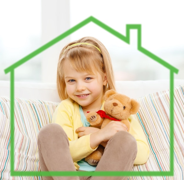 Foto concepto de infancia, ecología, gente y felicidad - niña sonriente con oso de peluche sentado en un sofá en casa detrás del símbolo de la casa verde