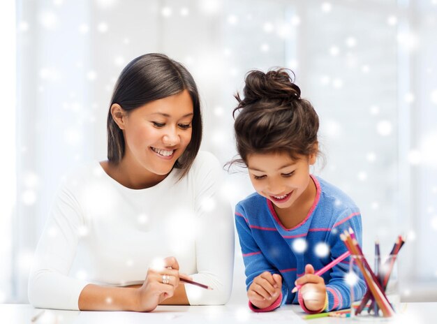 concepto de infancia, familia, educación y personas - niña sonriente y madre o maestra dibujando con lápices de colores en el interior