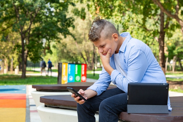 Concepto de infancia, realidad aumentada, tecnología y personas: niño con cara de desconcierto mira al teléfono inteligente al aire libre en verano