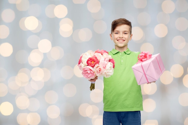 concepto de infancia, vacaciones, regalos y personas - niño feliz sosteniendo un ramo de flores y una caja de regalo sobre fondo de luces