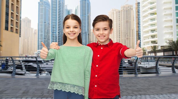 concepto de infancia, viajes, turismo, gestos y personas - niño y niña sonrientes felices abrazándose y mostrando los pulgares hacia arriba sobre el fondo de la calle de la ciudad de dubai