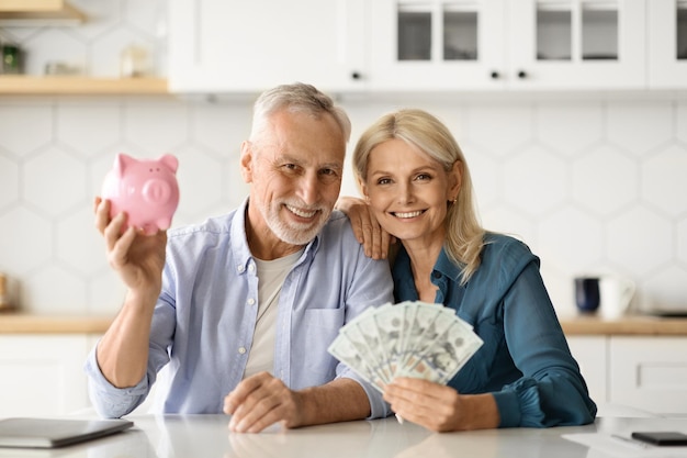 Foto concepto de inversiones sonriente pareja senior sosteniendo alcancía y dinero en dólares en casa