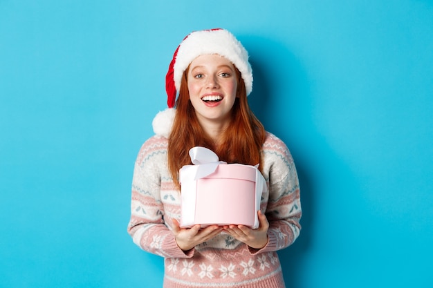 Concepto de invierno y celebración. Chica pelirroja feliz recibiendo regalo de Navidad y agradeciendo, abrazando la caja con regalos y sonriendo, de pie con gorro de Papá Noel sobre fondo azul.