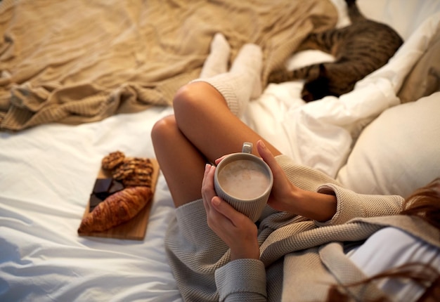 concepto de invierno, comodidad, ocio y personas - cerca de una mujer joven con una taza de café o cacao y dulces en la cama en casa