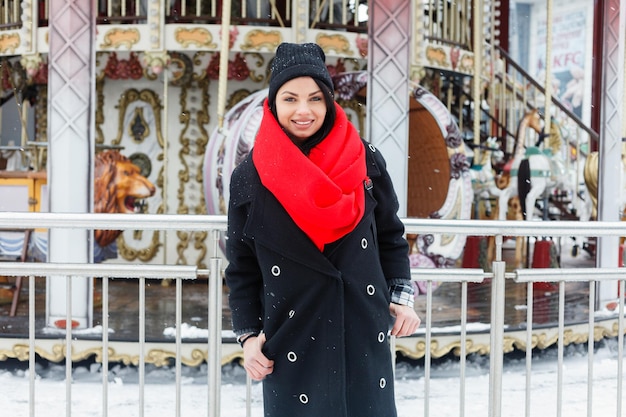 Concepto de invierno, navidad, estilo de vida, vacaciones y moda - mujer sonriente bastante joven en abrigo negro y bufanda roja posando en la ciudad de invierno. atractiva morena fondo de tiovivo francés