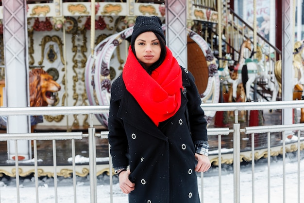Concepto de invierno, navidad, estilo de vida, vacaciones y moda - mujer sonriente bastante joven en abrigo negro y bufanda roja posando en la ciudad de invierno. atractiva morena fondo de tiovivo francés