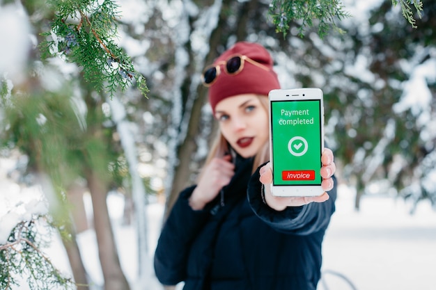 Concepto de invierno, navidad, tecnologías, personas, estilo de vida y belleza: mensaje de pago completado en la pantalla de un teléfono móvil. Mujer mostrando su teléfono móvil. Mujer de invierno que sopla nieve en un parque.