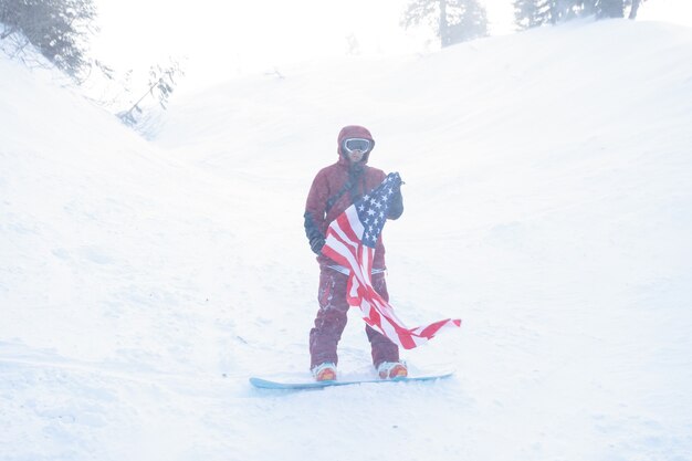 Concepto de invierno, ocio, deporte y personas: el snowboarder se sienta en lo alto de las montañas en el borde de la pendiente y mira a lo lejos. snowboarder, tenencia, bandera americana