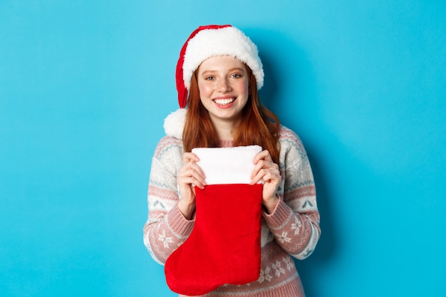 Concepto de invierno y vacaciones. Chica alegre recibiendo regalos en calcetines de Navidad, con gorro de Papá Noel y sonriendo feliz, de pie sobre fondo azul.