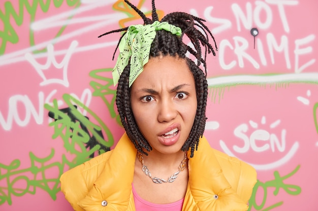 Foto concepto de jóvenes millennials de arte moderno. disgustada adolescente afroamericana tiene dreadlock sonríe cara posa contra la pared de graffiti está descontento