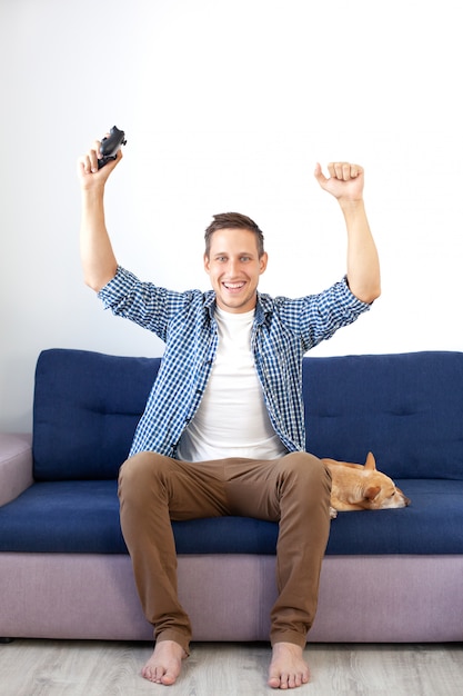 El concepto del juego. El chico está jugando un videojuego con un joystick en casa con un perro. Un hombre sonriente con camisa, sentado en el sofá, juega un videojuego con un joystick. Jugador. Juego de consola