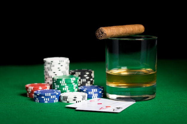 concepto de juego, fortuna y entretenimiento: cierre de fichas de casino, vaso de whisky, naipes y cigarros en la superficie de la mesa verde