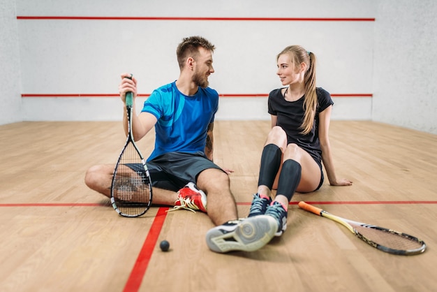 Concepto de juego de squash, raquetas con pelota, pareja joven sentada en el suelo después del entrenamiento activo